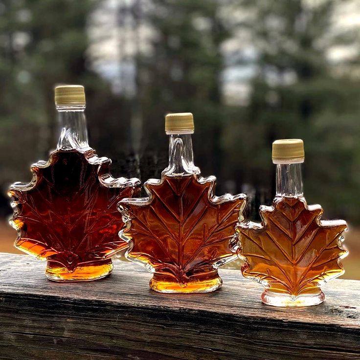 Three maple syrup bottles shaped like leaves on a wooden surface with a blurred natural background.